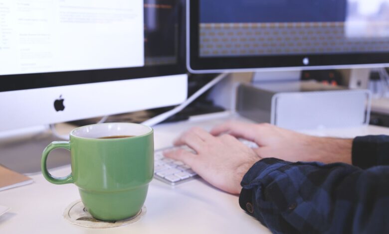 Person typing at a desk with coffee and computer screens. Perfect for business or technology themes.