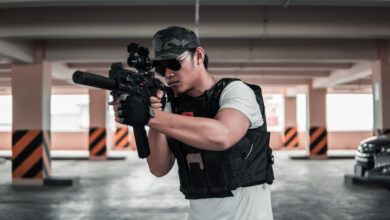 A man dressed in tactical gear aims a rifle in a shaded parking area, suggesting a training scenario.