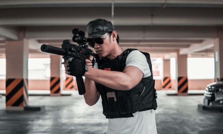 A man dressed in tactical gear aims a rifle in a shaded parking area, suggesting a training scenario.