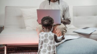 A father working on a laptop while his young daughter plays nearby, creating a family and work-life balance.