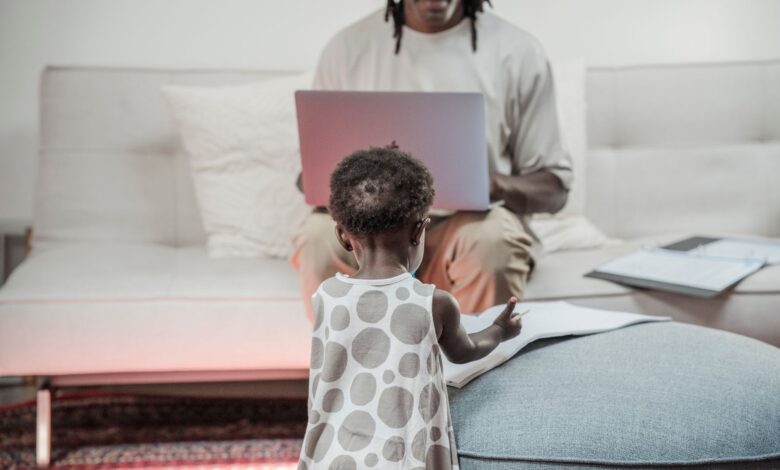 A father working on a laptop while his young daughter plays nearby, creating a family and work-life balance.