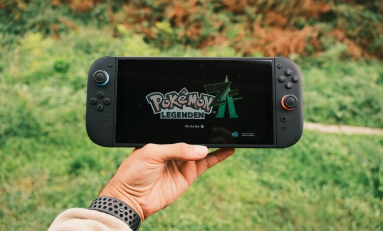 A hand holding a handheld gaming console displaying the Pokémon Legends game screen outdoors.