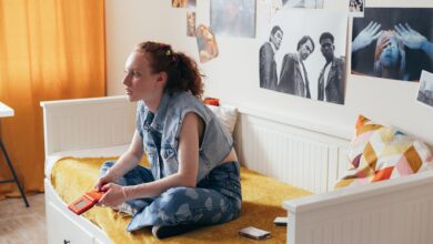 A woman in a denim vest sits on a bed holding a gamepad, surrounded by art posters.