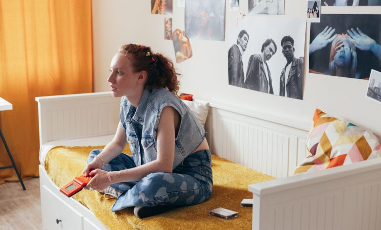 A woman in a denim vest sits on a bed holding a gamepad, surrounded by art posters.