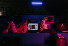 Teenagers play video games in a neon-lit setting with vintage monitors, capturing a retro aesthetic.