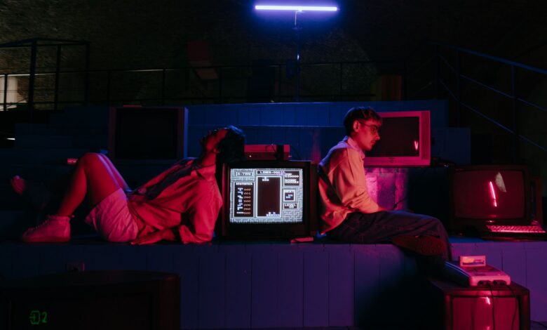 Teenagers play video games in a neon-lit setting with vintage monitors, capturing a retro aesthetic.