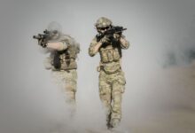 Two soldiers in camo uniforms with weapons advancing through a smoke-covered field, showcasing military action.