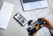 Person using vintage camera with smartphone and laptop in creative setup.