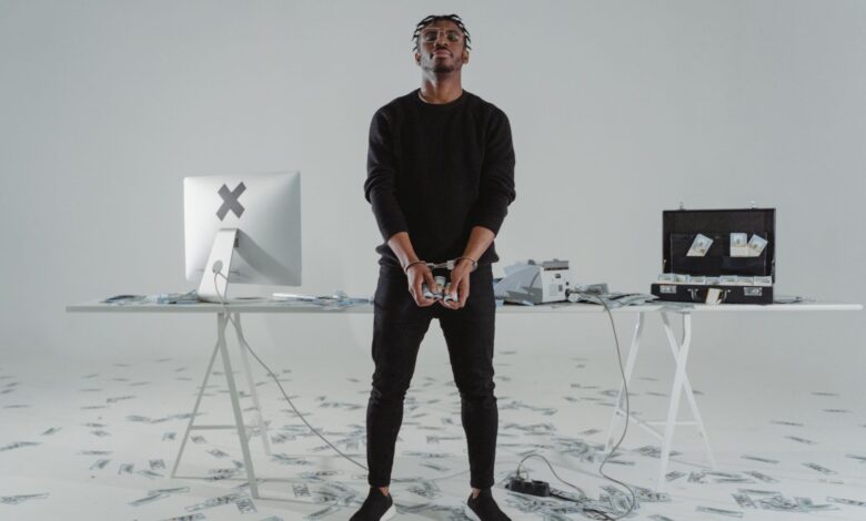 A handcuffed man stands in a room with scattered money, a computer, and a briefcase.
