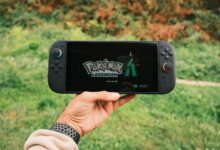 A hand holding a handheld gaming console displaying the Pokémon Legends game screen outdoors.