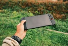 Close-up of a person's hand holding a black handheld gaming console against a green outdoor background.
