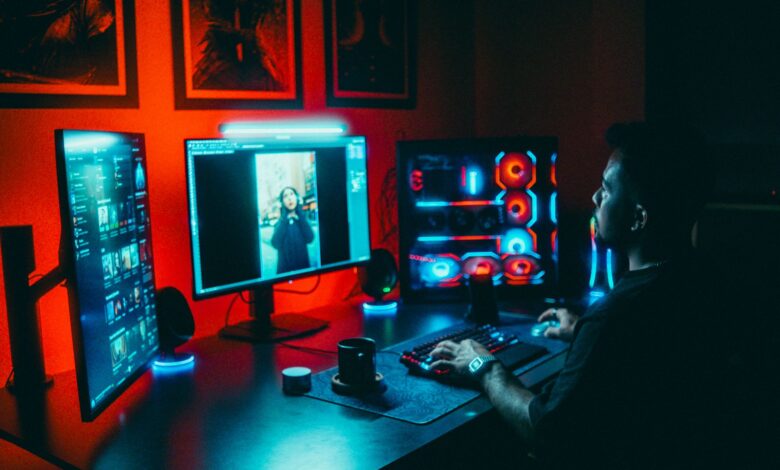 A young man working on a multi-screen computer setup in a dark, neon-lit room.