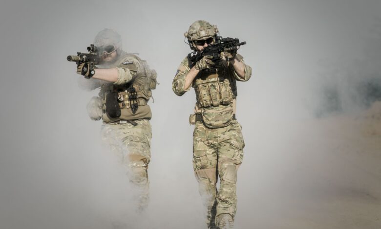 Two soldiers in camo uniforms with weapons advancing through a smoke-covered field, showcasing military action.
