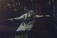 A figure in a whimsical costume poses eerily amidst a foggy, dark forest.