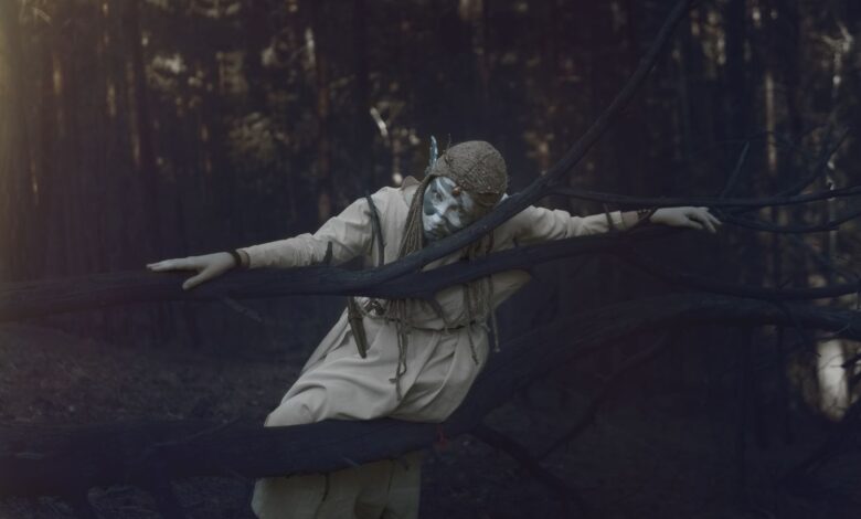 A figure in a whimsical costume poses eerily amidst a foggy, dark forest.