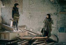 Two people exploring a desolate, abandoned building in post-apocalyptic attire.