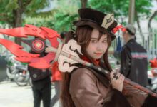 A female cosplayer dressed as Hu Tao from Genshin Impact poses outdoors with a detailed costume and weapon.