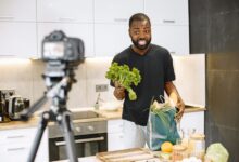 A man vlogging in a kitchen with fresh vegetables and recording equipment, showcasing a cooking segment.