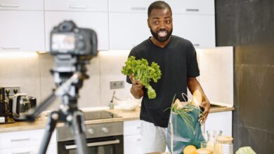 A man vlogging in a kitchen with fresh vegetables and recording equipment, showcasing a cooking segment.