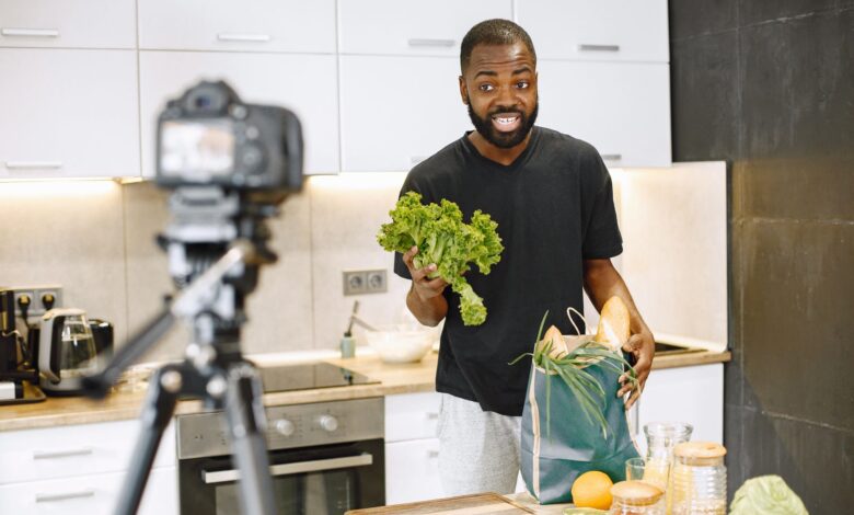 A man vlogging in a kitchen with fresh vegetables and recording equipment, showcasing a cooking segment.