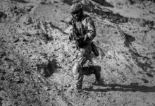 A soldier runs down a sandy hill in full combat gear under harsh desert sun.
