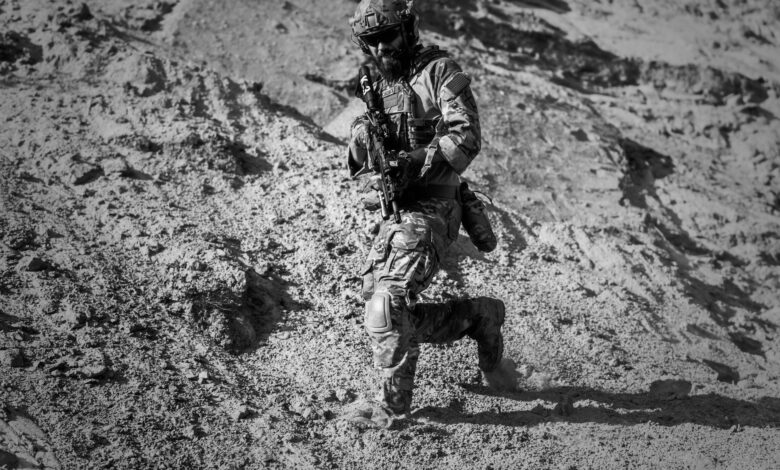 A soldier runs down a sandy hill in full combat gear under harsh desert sun.