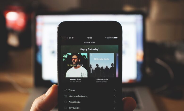 A person holding a smartphone with music streaming app in front of a laptop screen indoors.