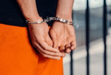 Close-up of person in handcuffs wearing orange attire behind prison bars, symbolizing arrest.