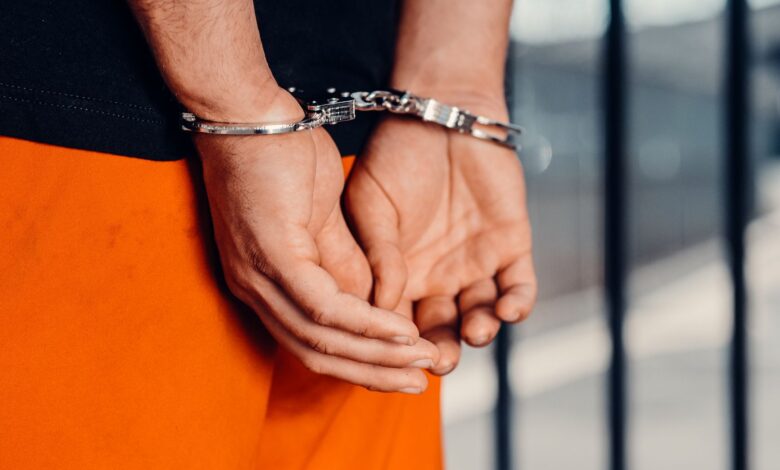 Close-up of person in handcuffs wearing orange attire behind prison bars, symbolizing arrest.