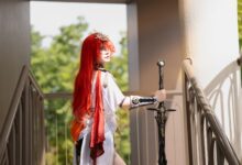 Cosplayer with striking red hair holds a sword on a sunlit stairway, dressed in elaborate costume.