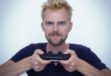 Man with short blonde hair fiercely focused while gaming with a controller against white background.