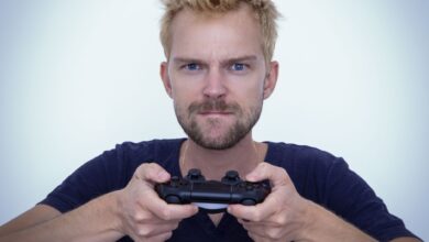 Man with short blonde hair fiercely focused while gaming with a controller against white background.