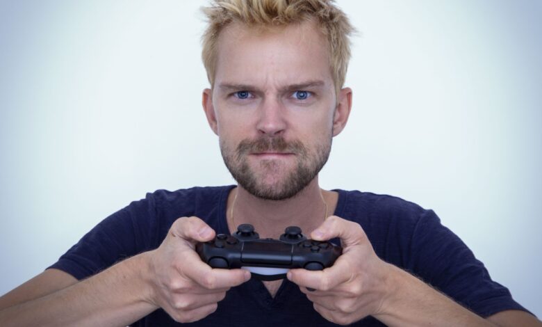 Man with short blonde hair fiercely focused while gaming with a controller against white background.