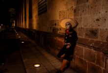 A man in Mexican costume with a painted face leans against a wall at night during a cultural festival.