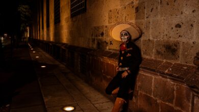 A man in Mexican costume with a painted face leans against a wall at night during a cultural festival.