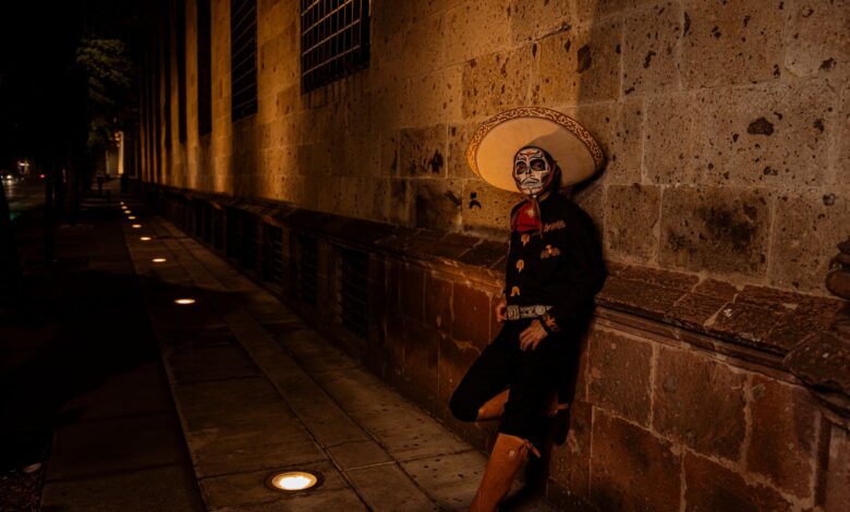 A man in Mexican costume with a painted face leans against a wall at night during a cultural festival.