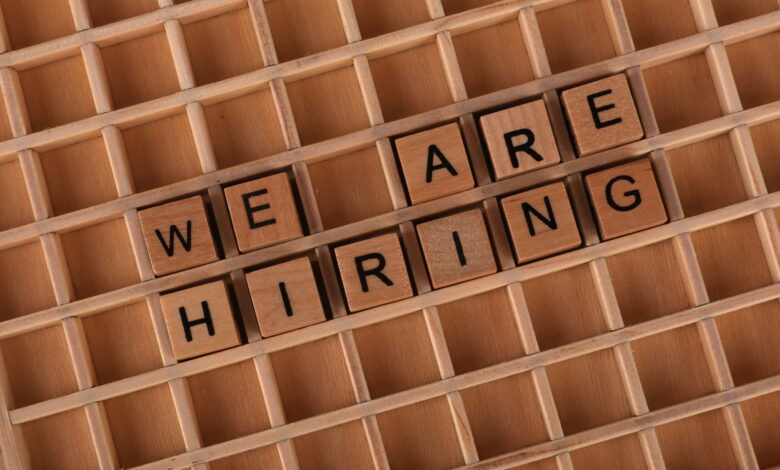 Wooden letter tiles arranged to spell 'We Are Hiring' on a wooden grid background.