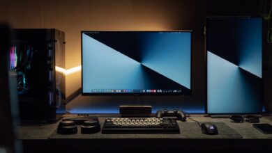 A dimly lit modern computer desk setup featuring dual monitors, a keyboard, and gaming accessories.