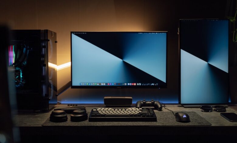 A dimly lit modern computer desk setup featuring dual monitors, a keyboard, and gaming accessories.