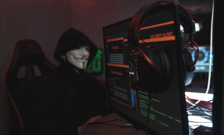 A person wearing a Guy Fawkes mask engaged in hacking activities on a computer in a dimly lit room.