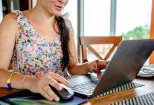A woman using a laptop for remote work indoors, featuring a floral dress and henna design.
