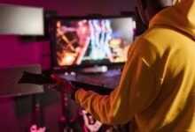 Man in yellow hoodie playing a music video game with a guitar controller indoors.