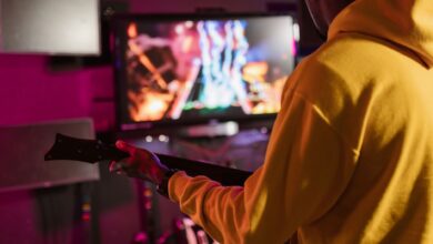 Man in yellow hoodie playing a music video game with a guitar controller indoors.
