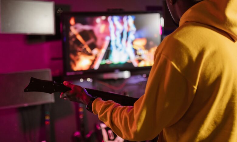 Man in yellow hoodie playing a music video game with a guitar controller indoors.