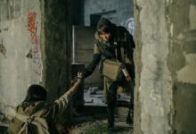 A man helps a woman in a desolate, post-apocalyptic urban environment.