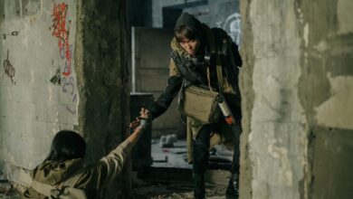 A man helps a woman in a desolate, post-apocalyptic urban environment.