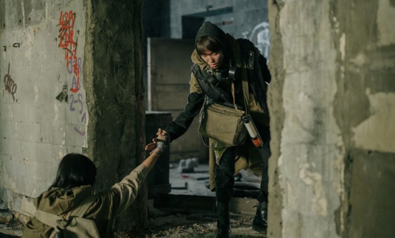 A man helps a woman in a desolate, post-apocalyptic urban environment.