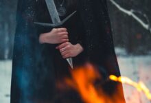 A cloaked figure holds a sword amidst snow and flames, evoking mystery.