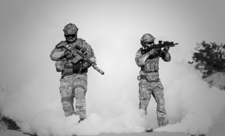 Black and white image of soldiers in a tactical operation amidst smoke and sand.