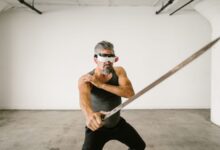 Adult man wearing VR headset practicing sword techniques in empty indoor space.